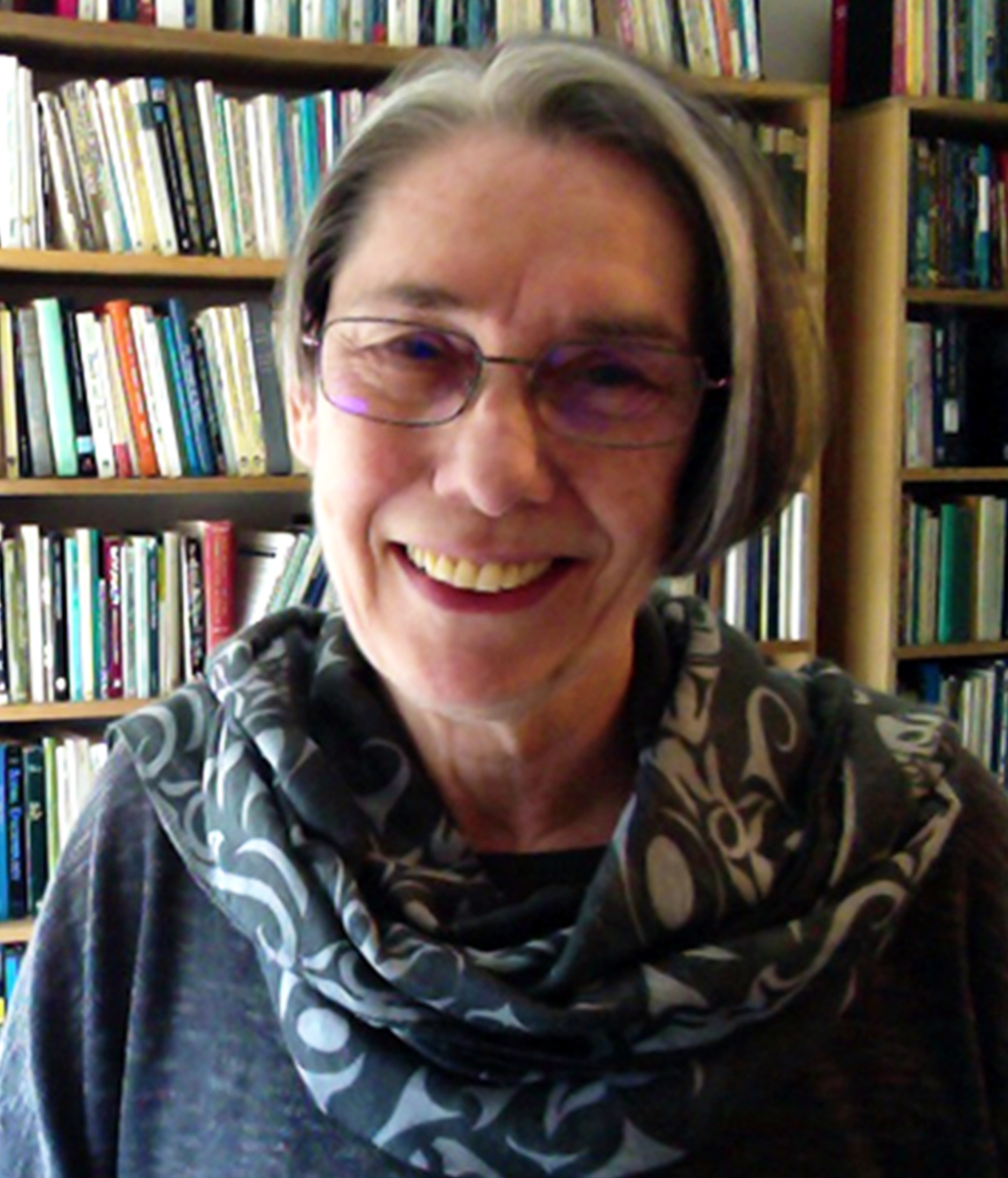 A white woman with gray hair and glasses, wearing a blue outfit, is in front of a bookcase and smiles at the camera.
