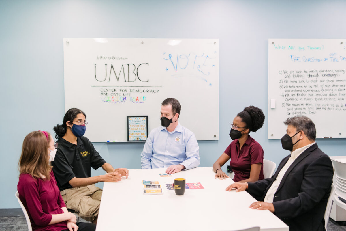 Two professors and three students sit around a white table talking. civic engagement. A dry erase board in the background reads "UMBC" and "VOTE."