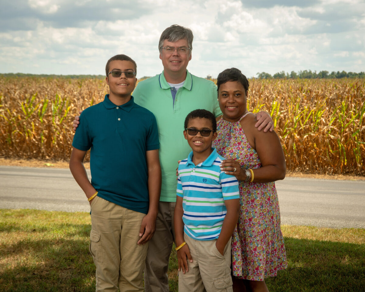 A family of four stands together in front of a field of corn on a sunny day.