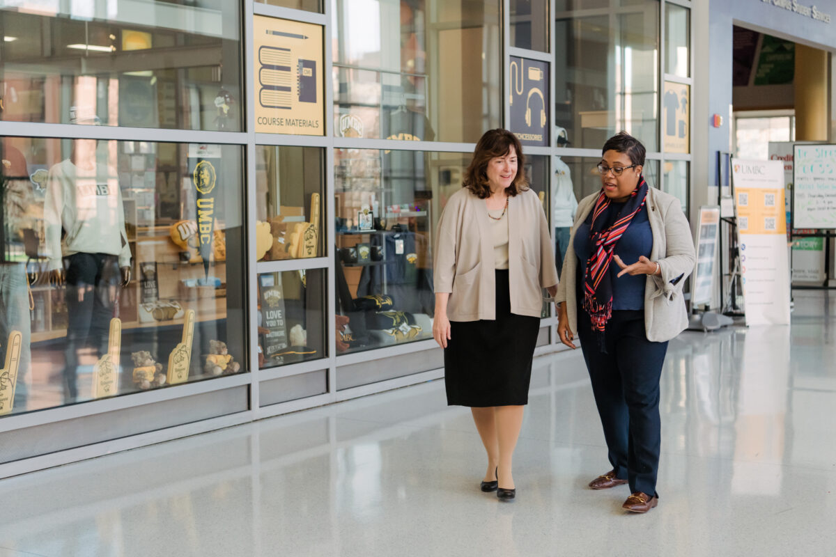 Two adults in business clothing walk and chat in a hallway with a wall of windows in the background talk about federal programs