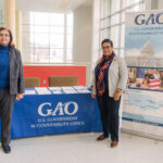 Two adults stand in front of a table with a blue table cloth with the letter GAO written in white a federal program.