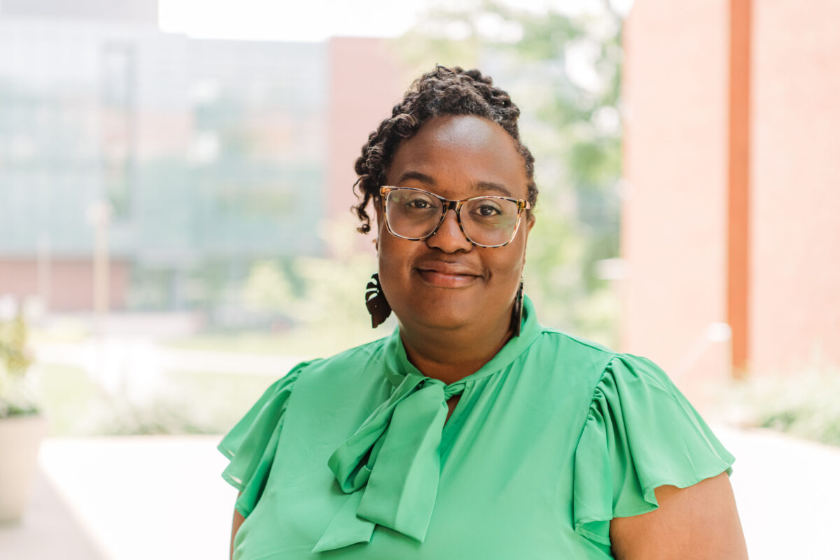 A person with dark brown wavy hair wearing a emerald blouse and glasses (NEH Fellowship recipient) stands outside with buildings in the background.