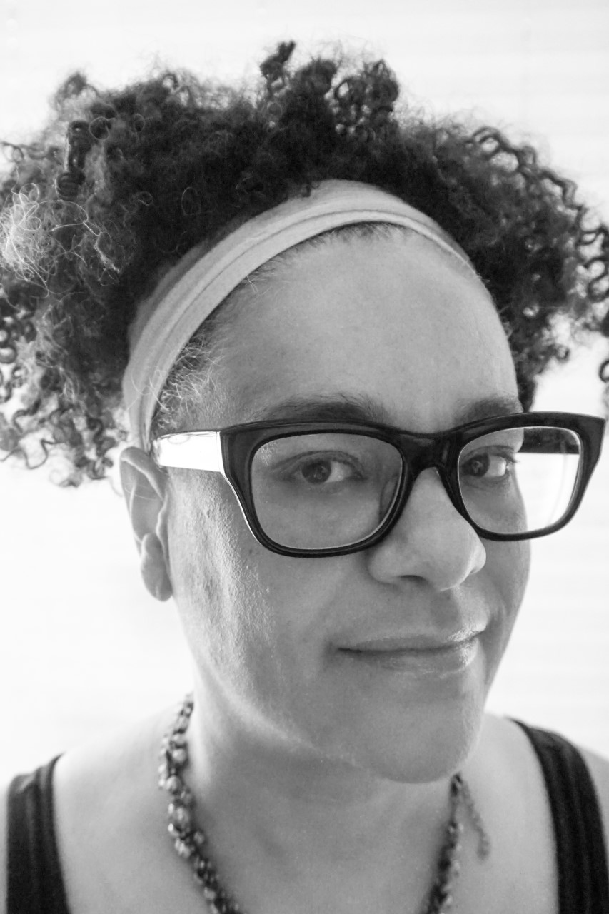 In a black and white photo, a Black woman wearing a hairband and glasses looks at the camera