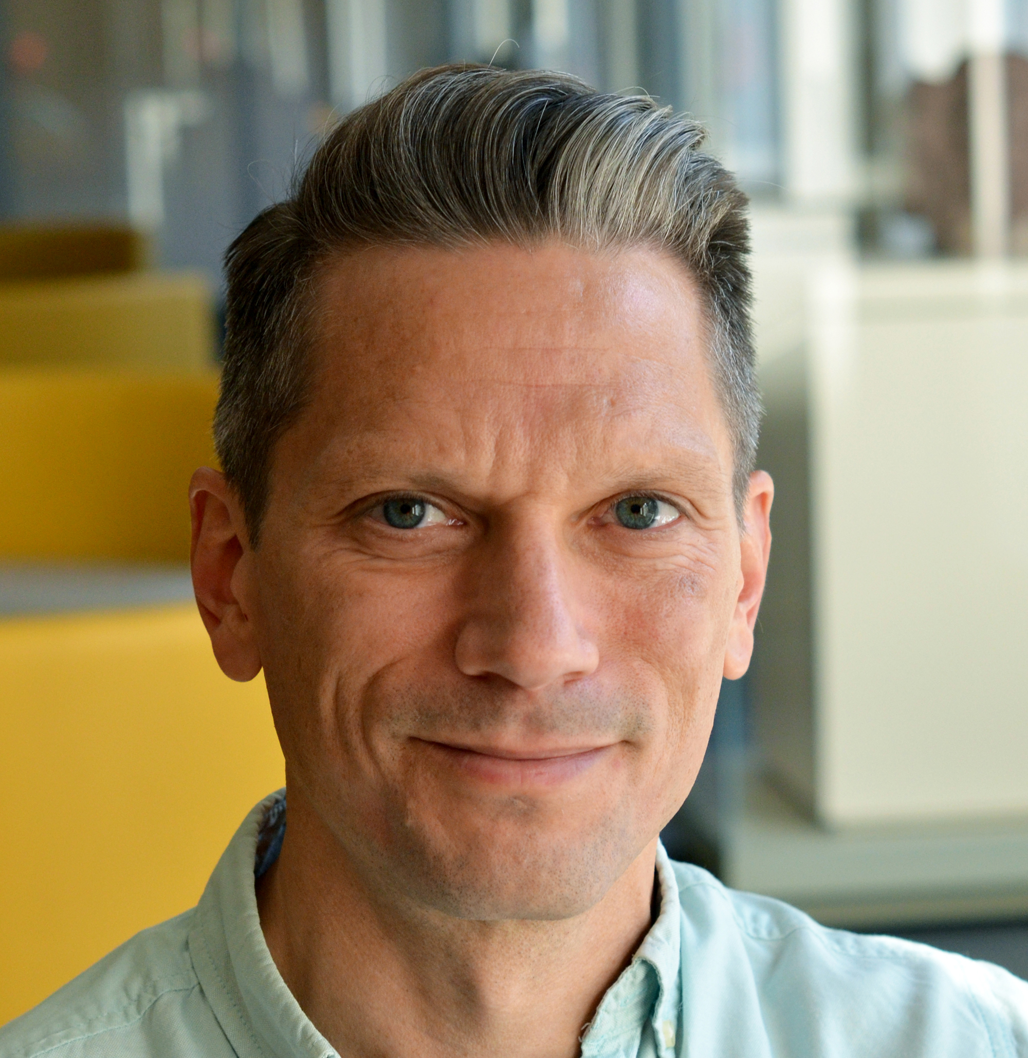 A white man with short gray hair and blue eyes is smiling at the camera. He has on a light blue collared shirt.