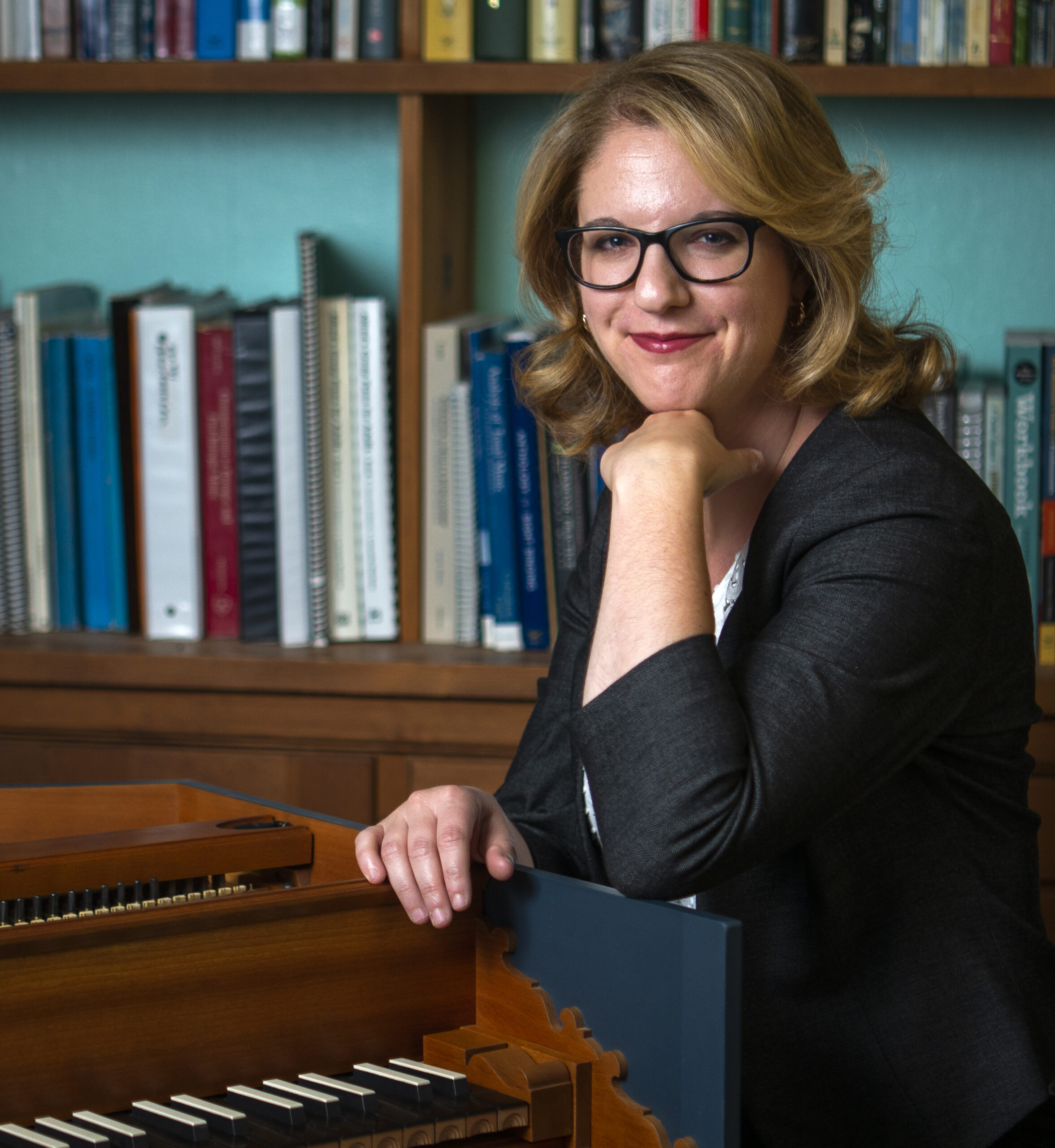 A white woman with a dark outfit and glasses smiles at the camera. She rests her arms on a harpsichord, and behind her is a bookcase.