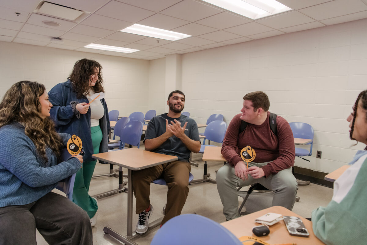 A group of students sit in a classroom, talking. Two hold signs reading "Paw Poll."