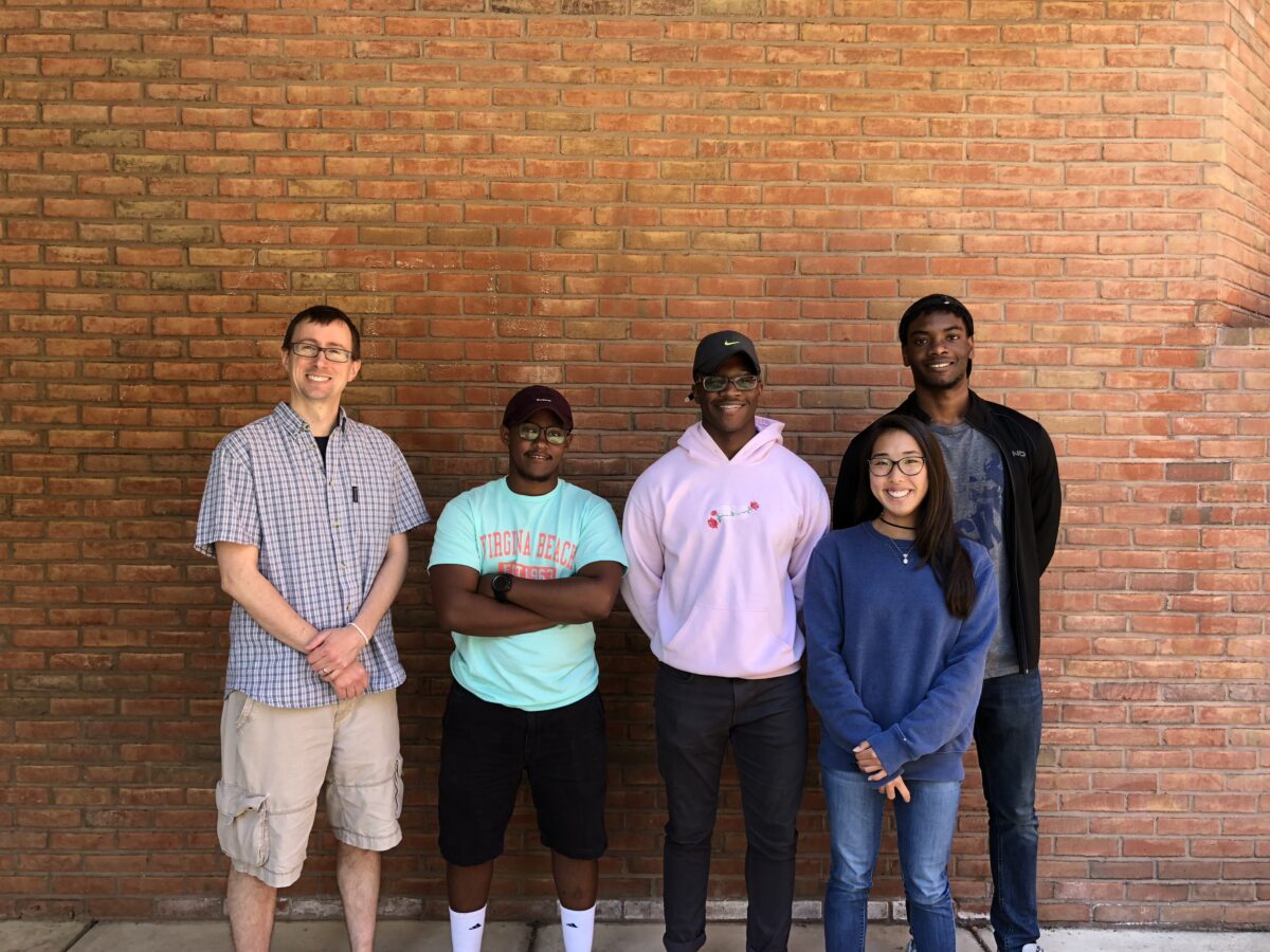 Five team members stand together with a brick wall in the background