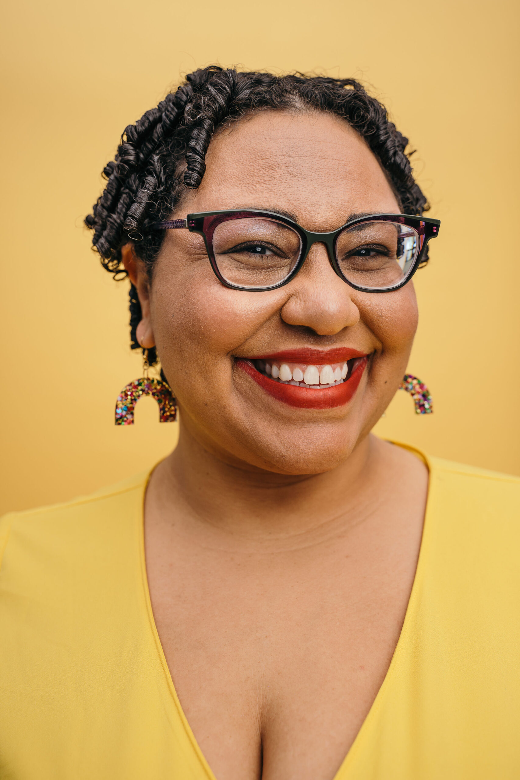 A Black women with short curly hair is smiling at the camera. She is wearing glasses with black frames and red liptstick.