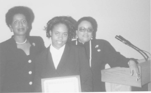 a black and white photo of three women standing at a podium, the woman in the center holds a certificate