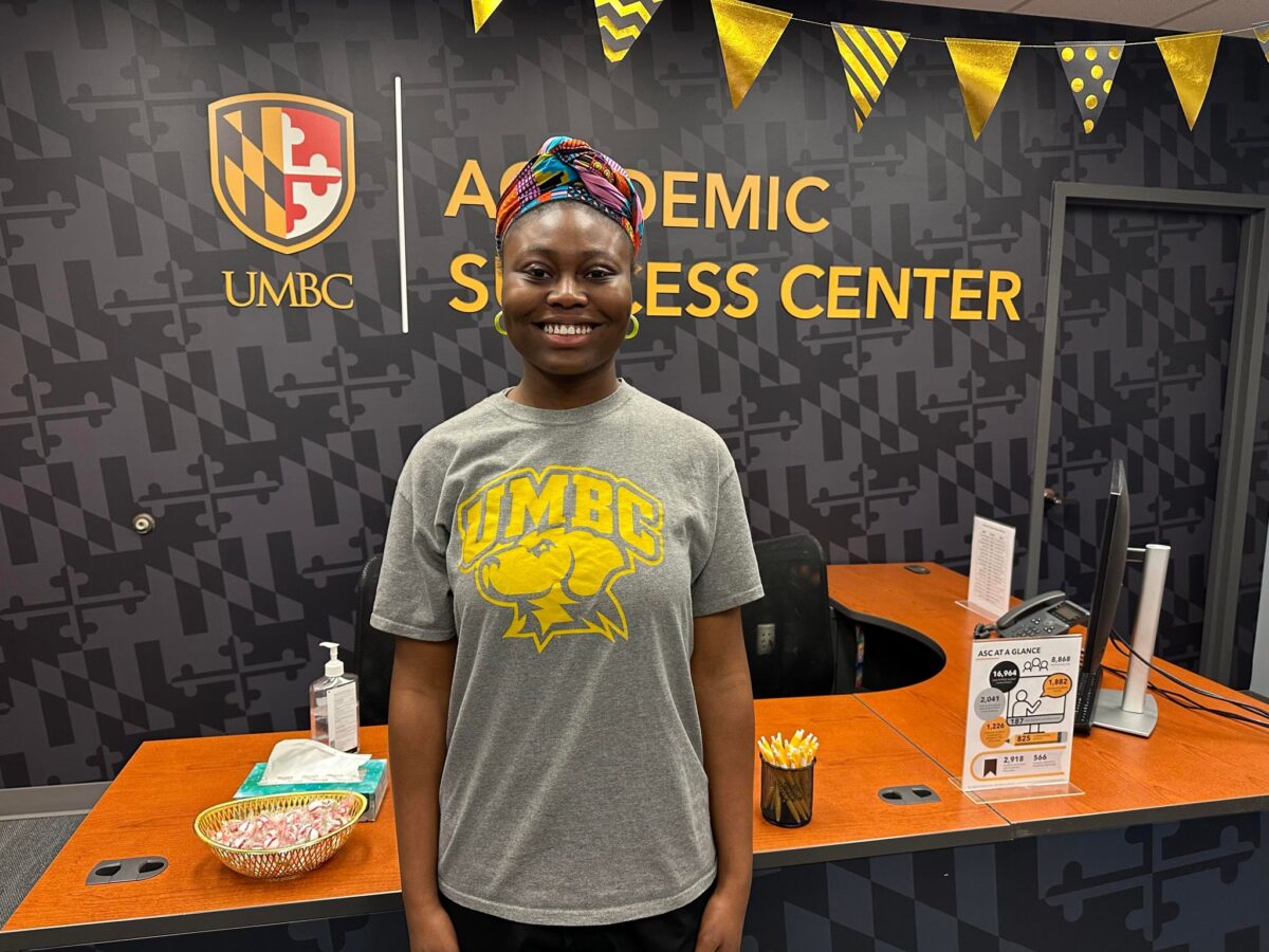A woman in a gray UMBC t-shirt stands in front of a desk where she acts as an academic peer advocate.