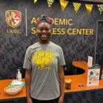 A woman in a gray UMBC t-shirt stands in front of a desk where she acts as an academic peer advocate.