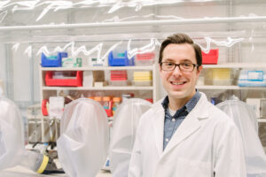 Man in white lab coat standing in front of a chemical hood with lots of colorful containers.