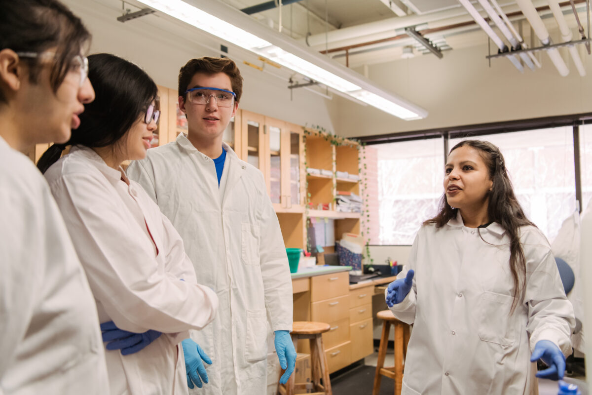 Four people in a laboratory, wearing lab coats and gloves. One stands to the right, speaking to the other three.