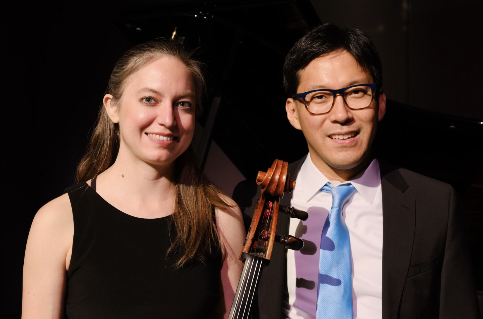 A woman wearing a black top and a man with glasses in a suit with a light blue tie smile at the camera. The neck of a cello is between them.