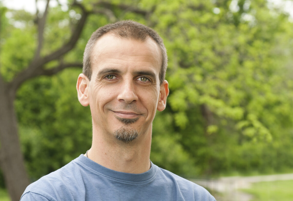 Heashot of man (antibiotic resistance researcher) in blue-gray t-shirt, greenery in background