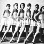 A black and white photo of a Black Vaudeville troupe of seven dancers posing in a row while sticking their leg out in unison.