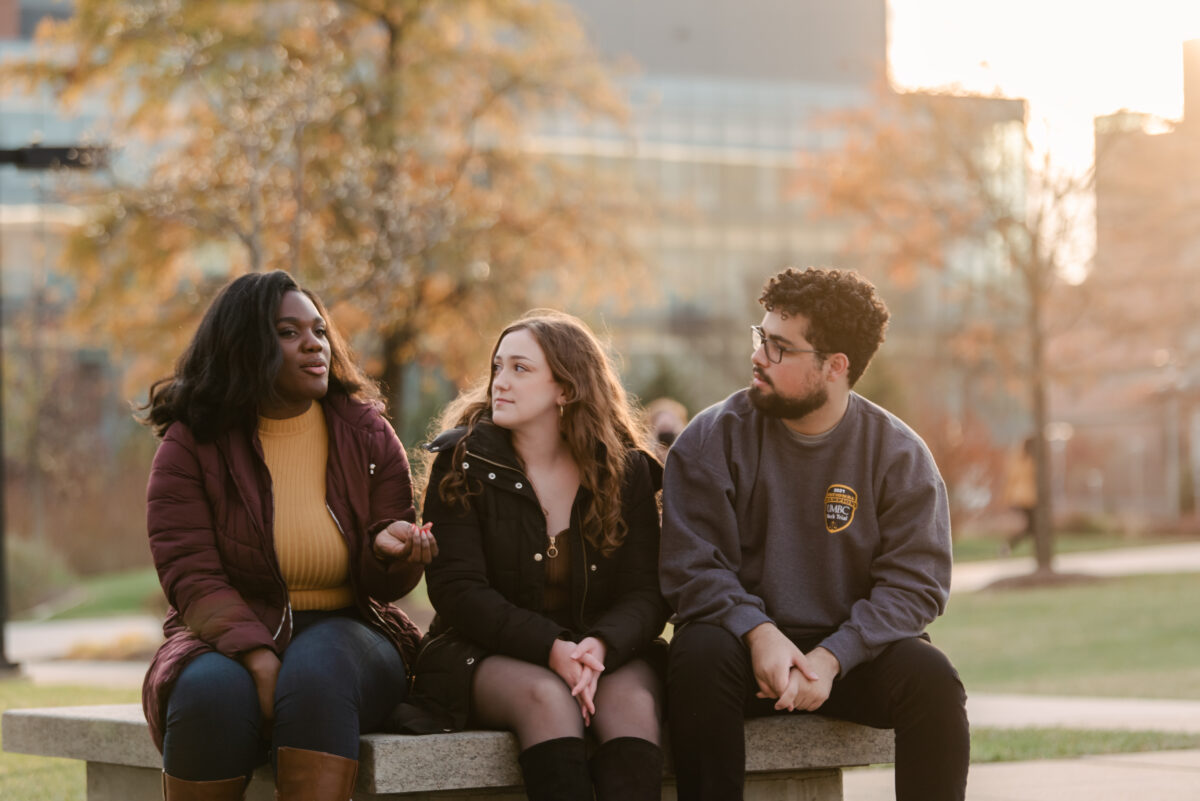 3 students of diverse race and genders, sitting closely on a bench outside, engaged in conversation