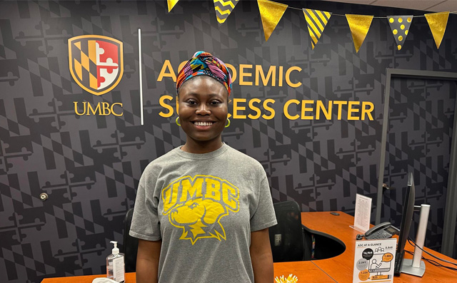A young Black woman in a t-shirt and a colorful head wrap smiles at the camera