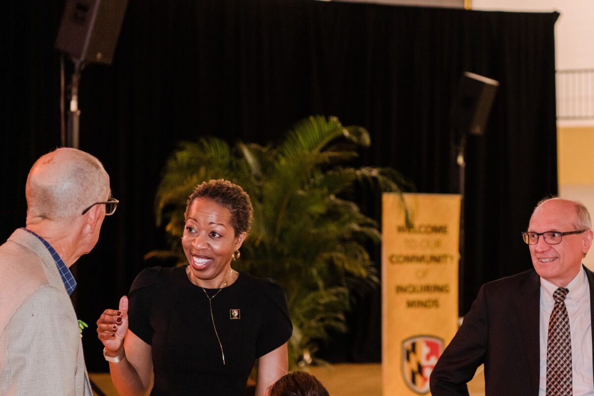 Woman in black dress speaks in an engaged way with a man in a gray suit. Another man in a suit smiles from a few steps away.