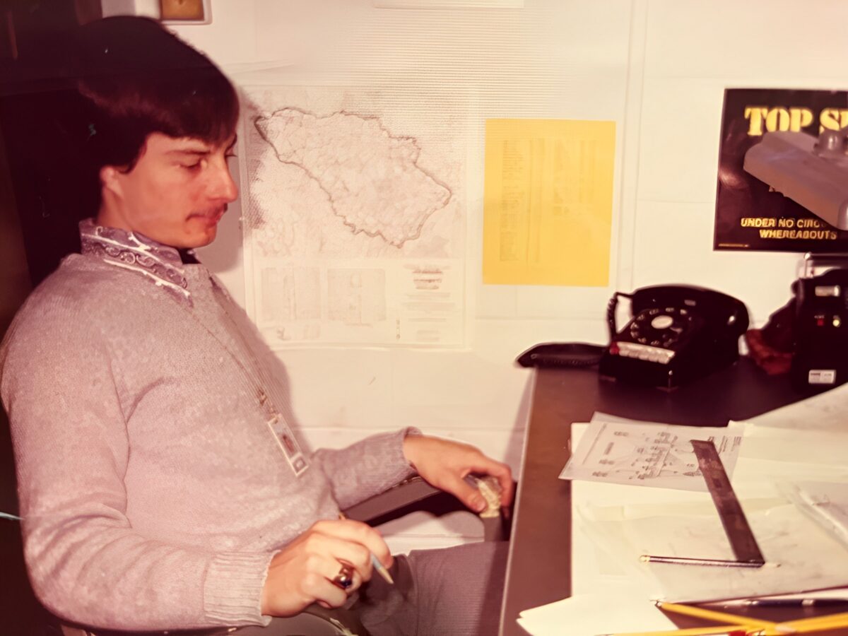In a retro photo, a young man working in nuclear national security sits at a desk doing math with a rotary phone and a map in front of him.