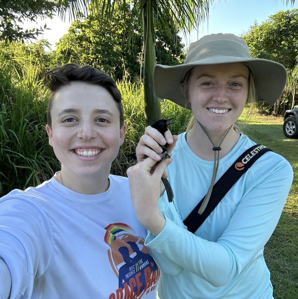 selfie of two student researchers outdoors in a tropical field site in field clothes, one holding a black bird