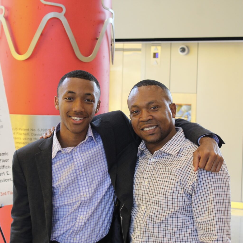 A father and son stand with their arms around each other.