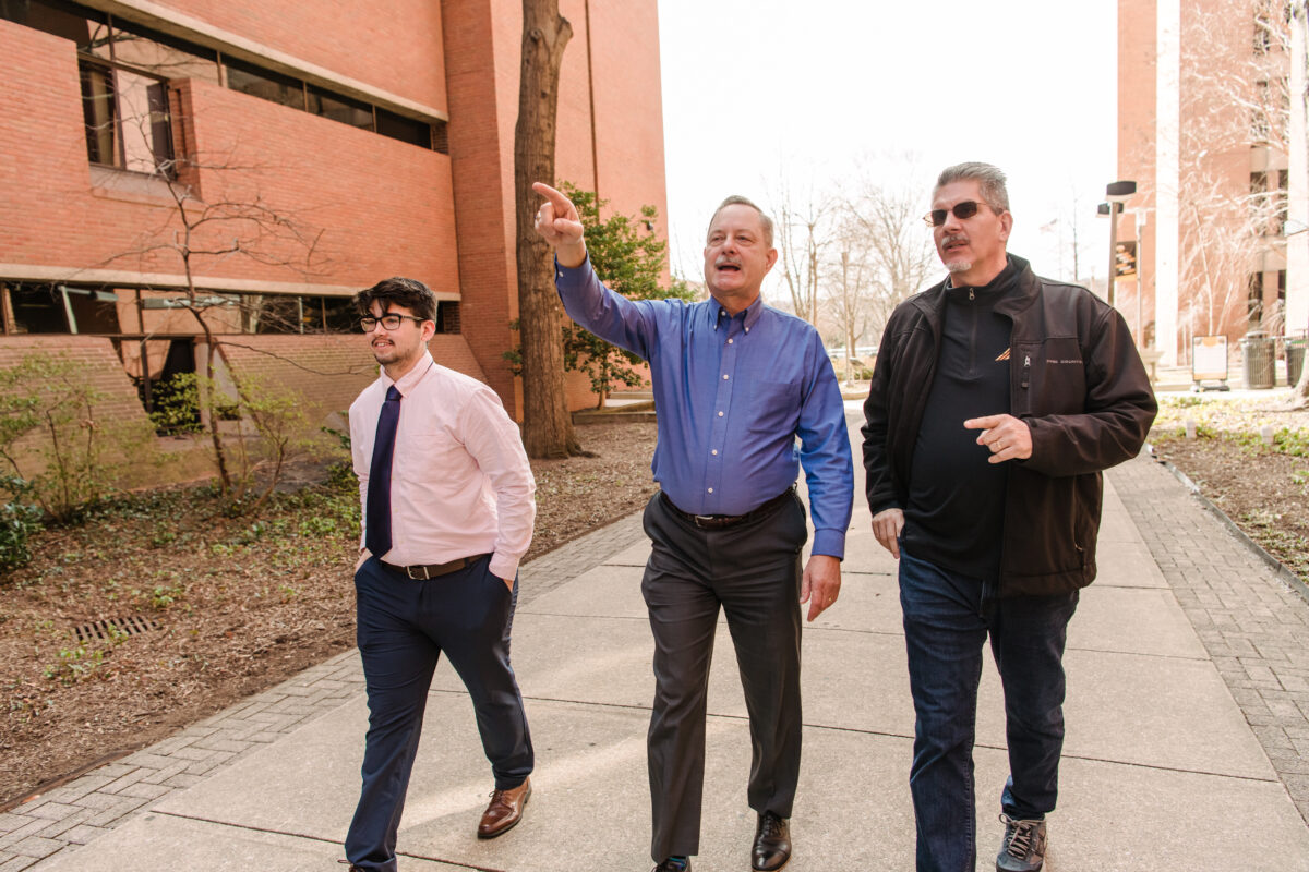 Three men walk down academic row, the middle man points to something in the distance