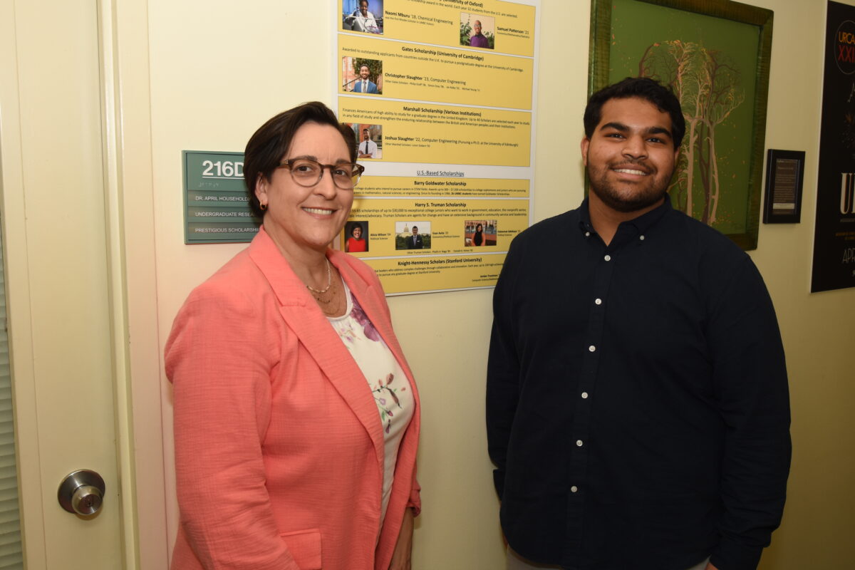 Faculty member and student pose in a hallway in front of a yellow poster with student photos on it.