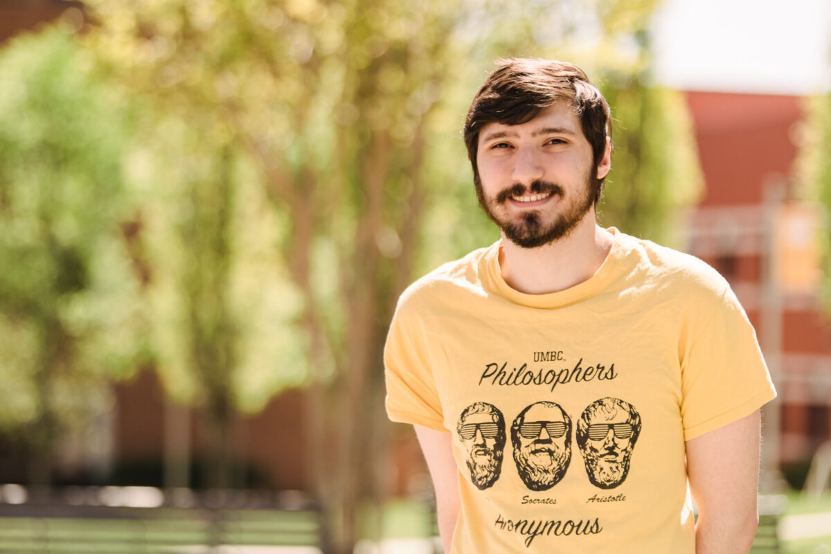 A science student with short brown hair, mustache, and beard wearing a yellow t-shirt about philosophy stands outside in front of a row of trees