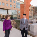 A woman in bright purple walks and talks next to a man in a suit jacket outside next to a sculpture. she is helping connect him to faculty funding and awards