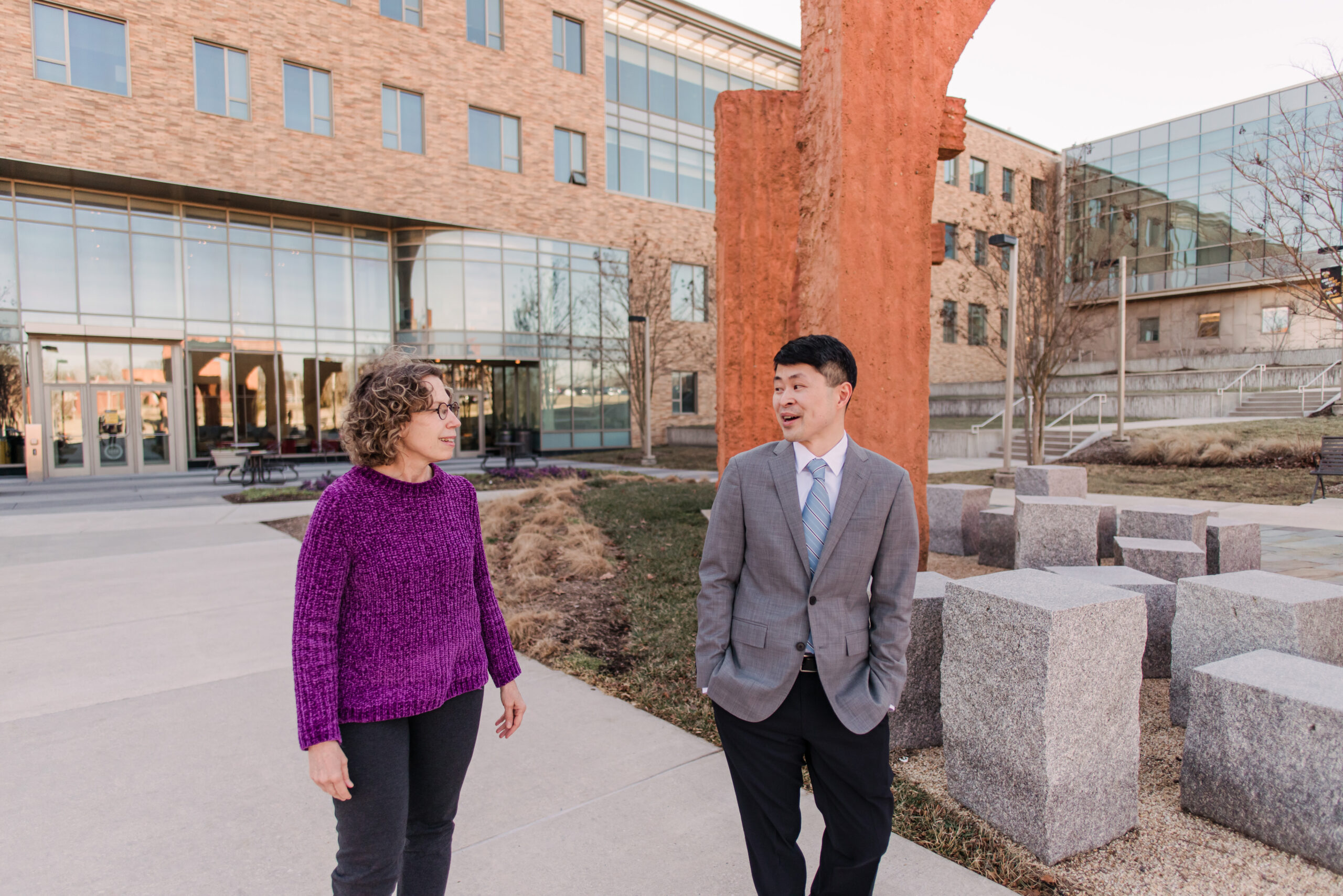 A woman in bright purple walks and talks next to a man in a suit jacket outside next to a sculpture. she is helping connect him to faculty funding and awards