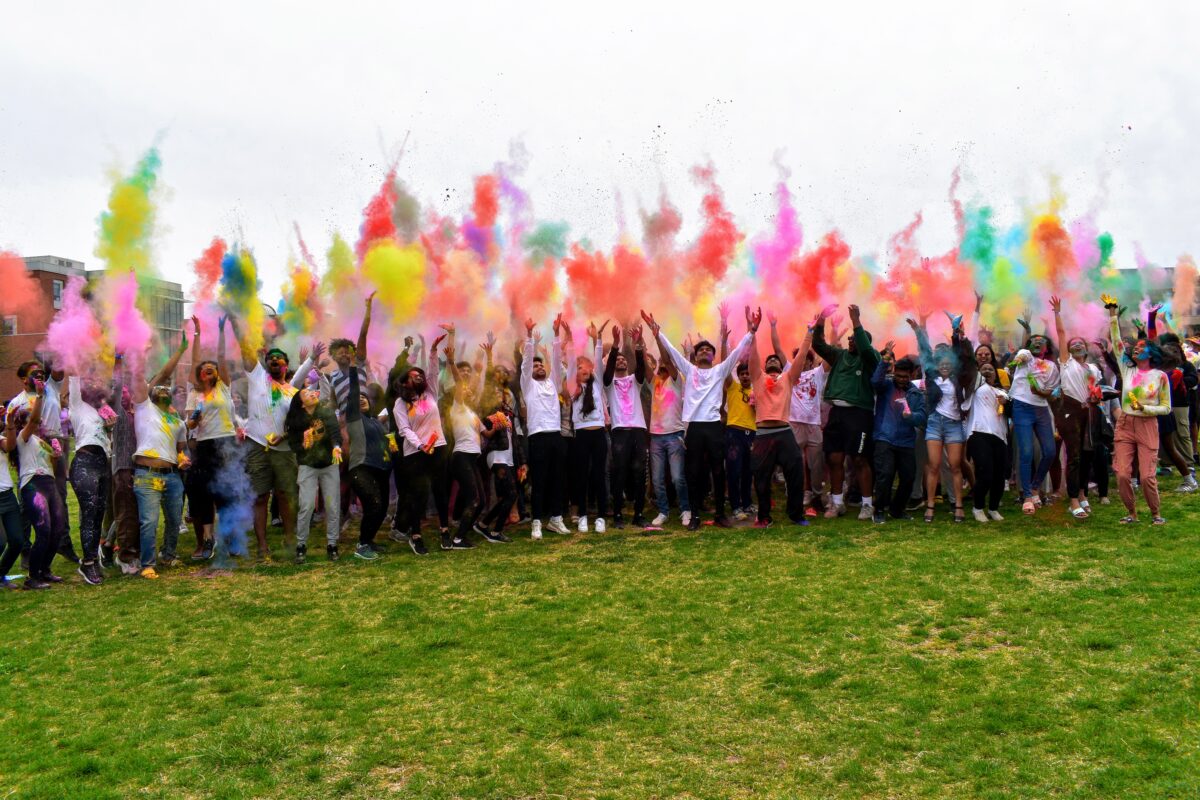 A community of people toss colorful powder into the sky, creating a line of colors
