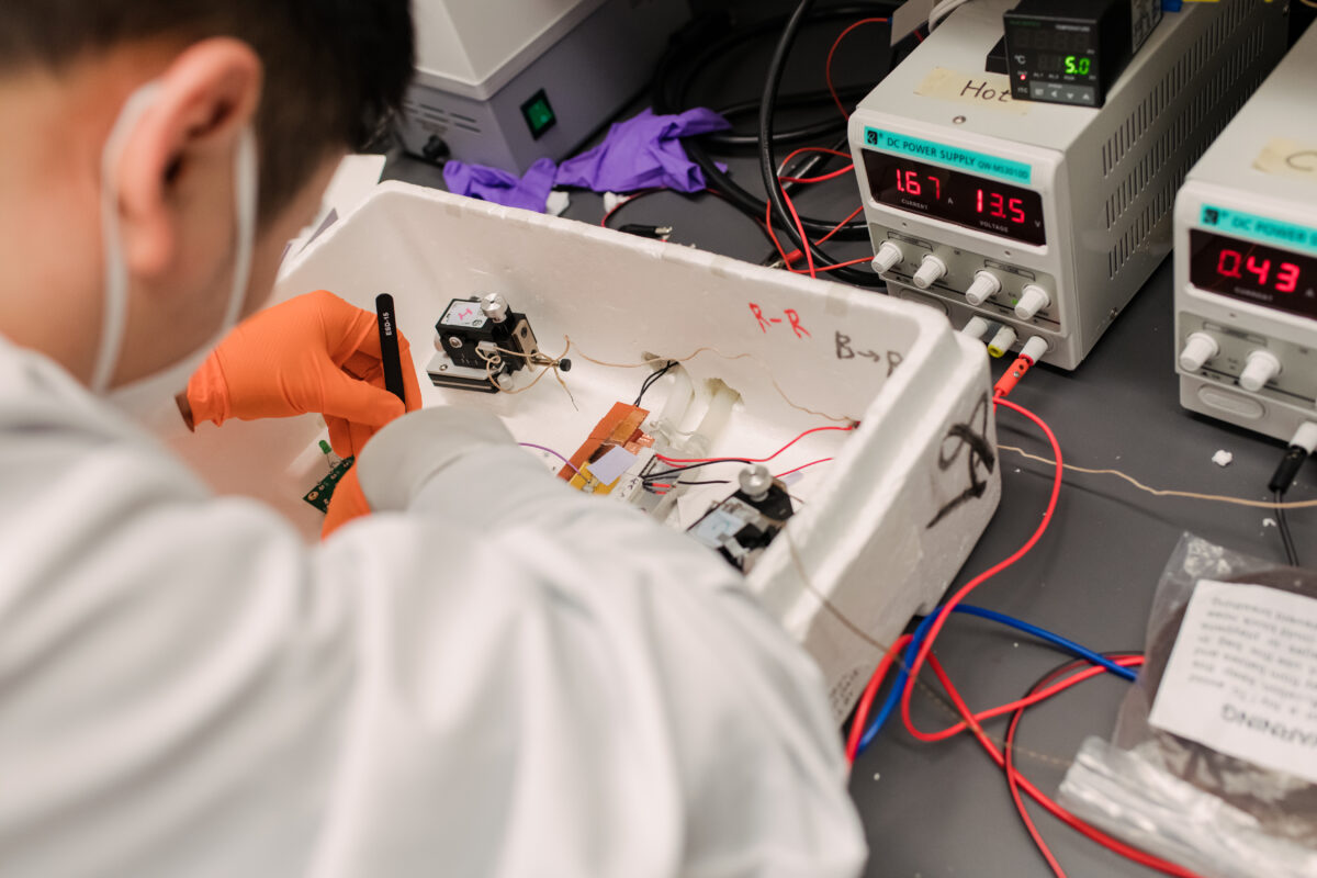 Person works on electronics in a lab, wearing a white coat, face covering, and bright orange gloves.