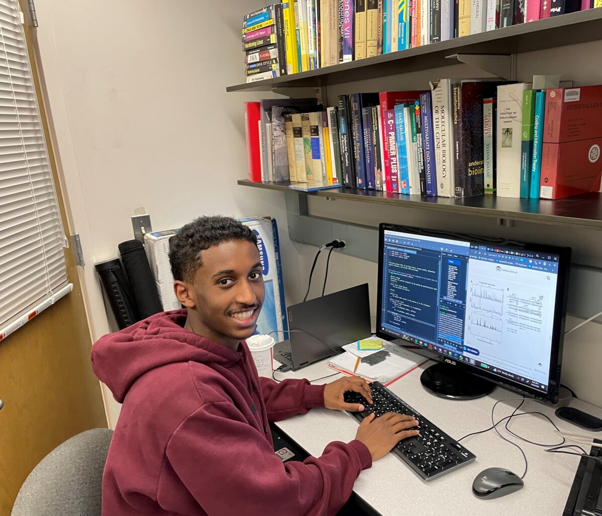 Student smiling while working on a desktop computer, with computer code and charts visible on the screen and two shelves of books in the background.