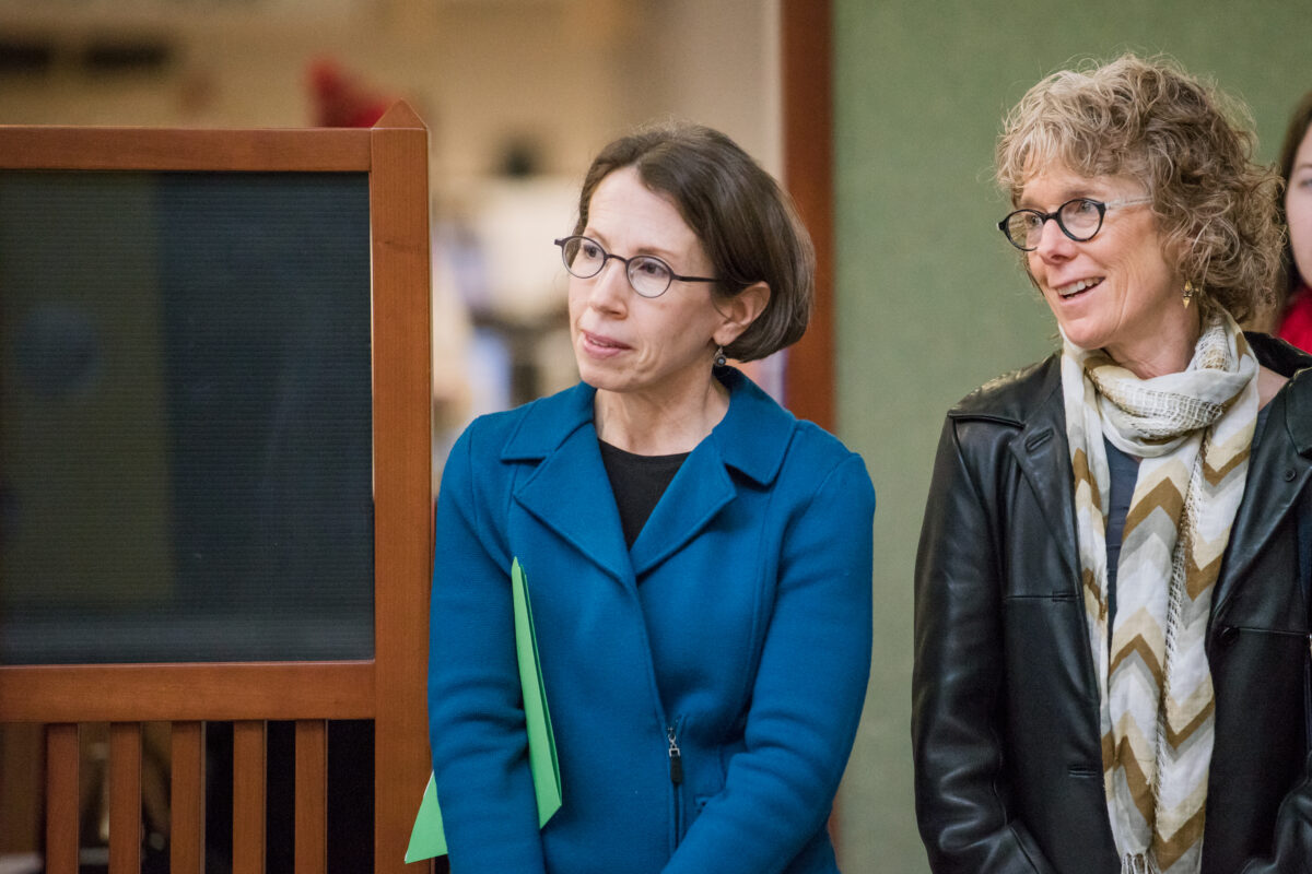 Two woman stand attentive with a black board behind them