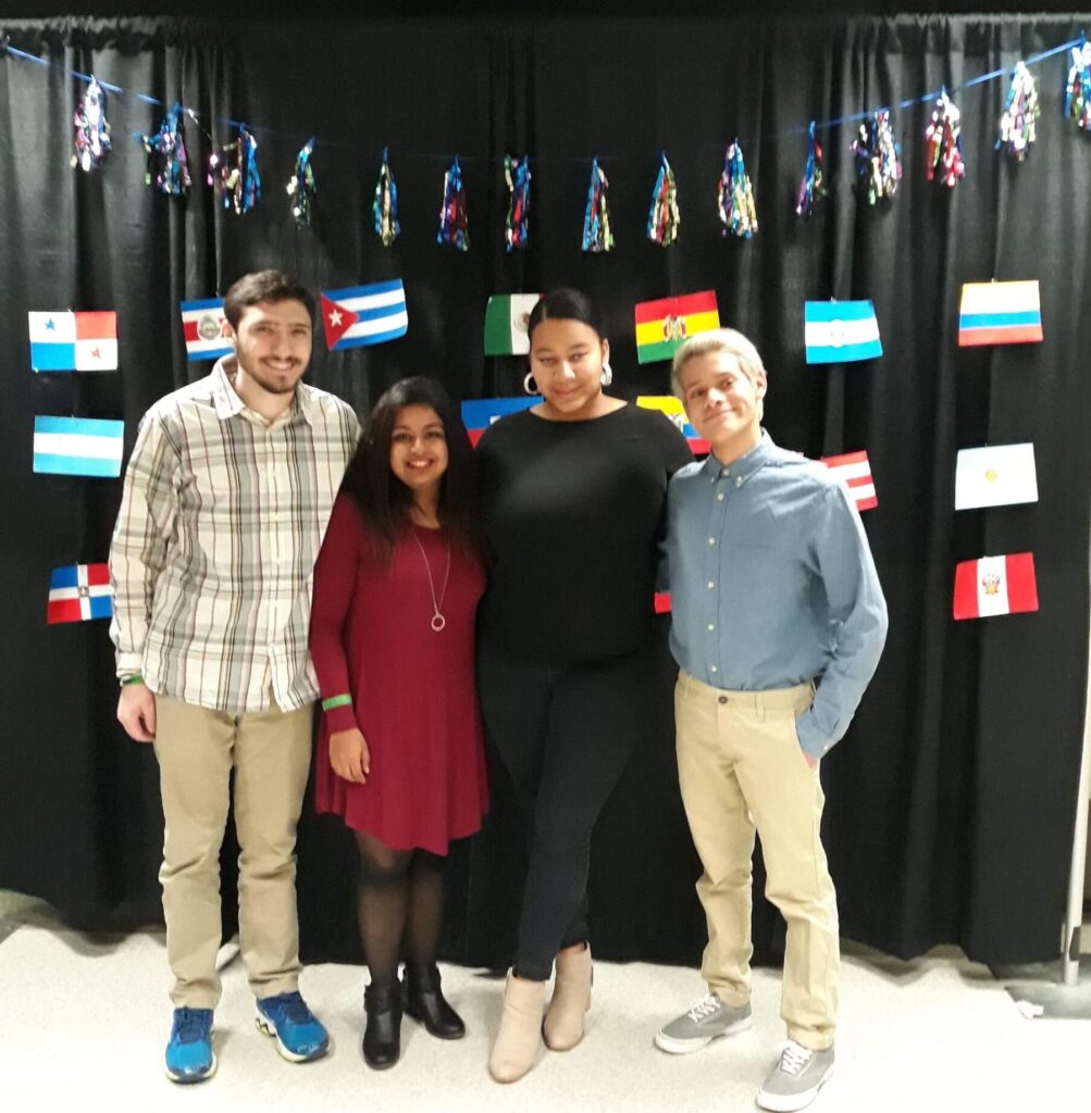 A group of science and humanities students stand arm in arm in front of a black curtain with pictures of flags from Latin American countries. council of majors
