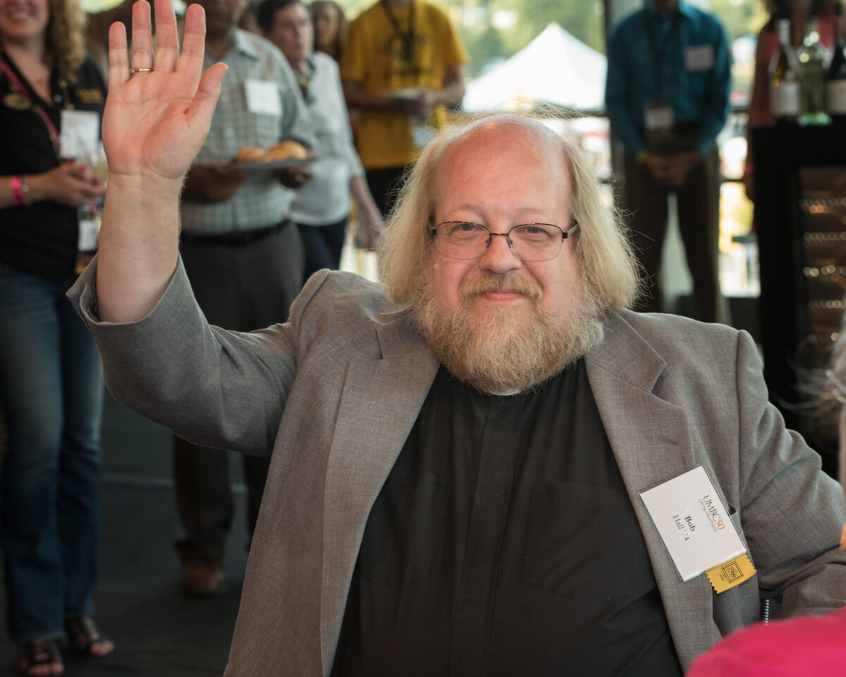 A man with long gray hair and matching beard and glasses waves. He was a philosophy major at UMBC.