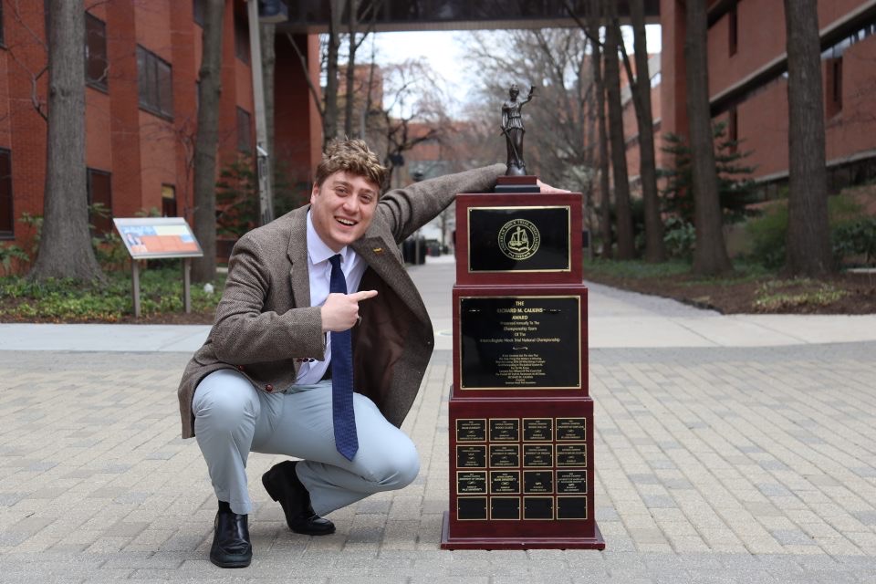 At the end of Academic Row, a student crouches next to a Mock Trial trophy about 3.5 feet fall; one hand rests on top of the trophy, the other is pointing at it.