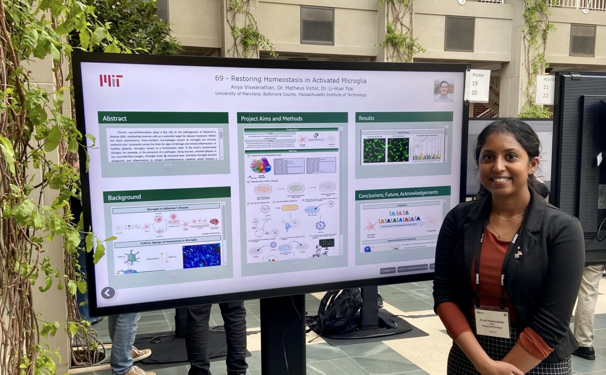 Student in professional attire and wearing a name badge lanyard stands next to a digital scientific poster in a high-ceilinged atrium.