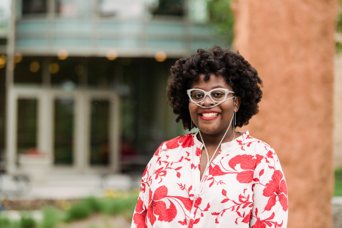 A student with light rimmed glasses and a red and white blouse stands outside with large cement arches behind her