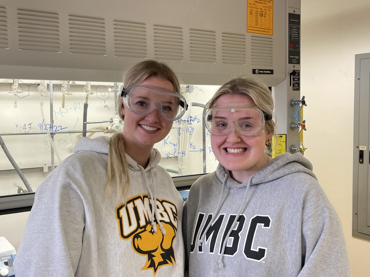 Two students smiling wearing UMBC sweatshirts while in a research lab, wearing safety goggles