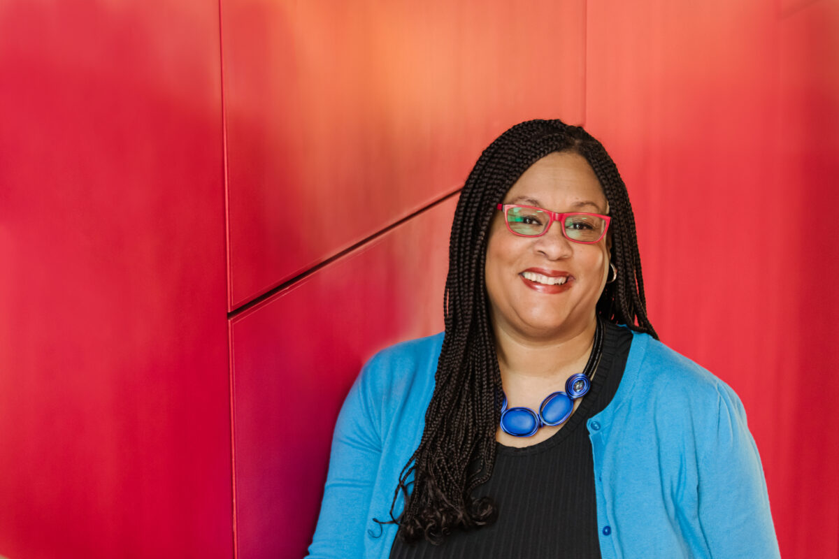 An adult with long, black braids wearing a black dress and a light blue sweater leans on a red wall. Black Vaudeville