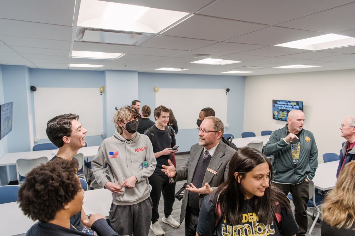 A crowd of people talk and laugh inside a room with tables, chairs and wall-mounted monitors.