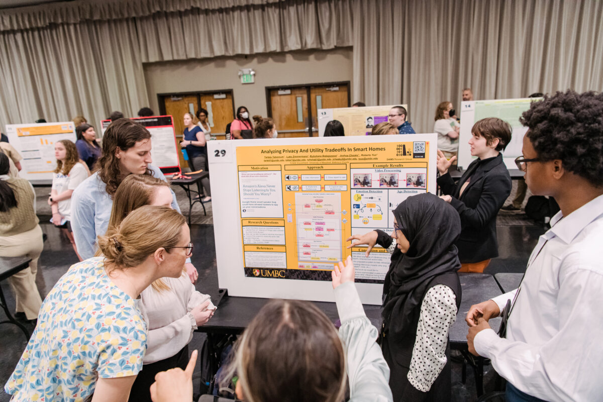 Student researchers explaining their research in front of poster board while conference attendees watch