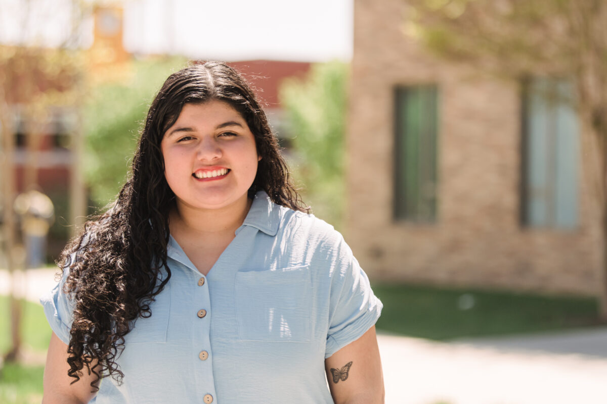 A first-generation college student with long brown wavy hair wearing a short sleeve light blue blouse stands outside on a sunny day in front of a brick building