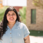 A first-gen immigrant student with long brown wavy hair wearing a short sleeve light blue blouse stands outside on a sunny day in front of a brick building