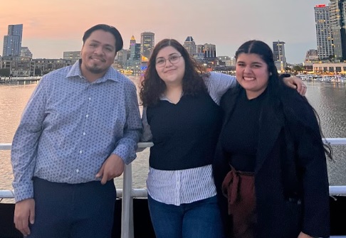 A Latinx group of three people stand close together with a city skyline in the background.