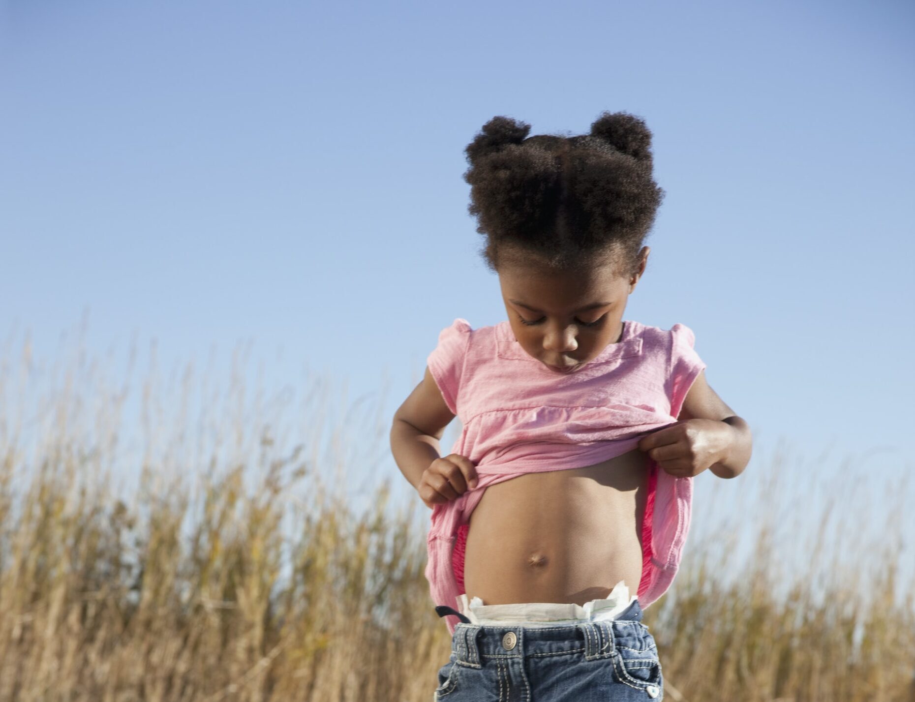 A little girl standing in a grassy field holds up her shirt to look at her belly button.