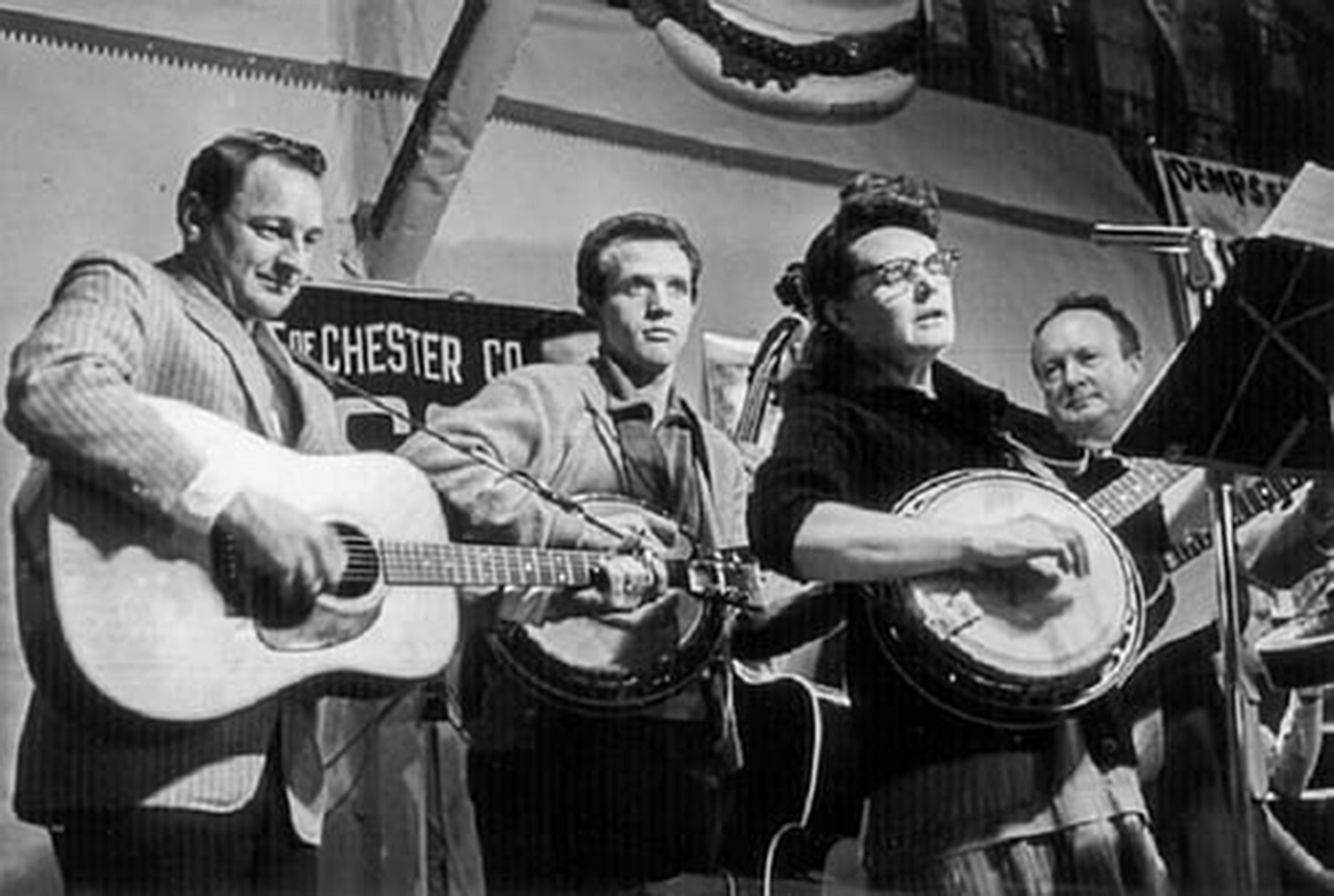 In an old black and white television image, a group of musicians play instruments