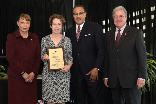 Four people stand and grin for a photo, a woman holds an award in her hands